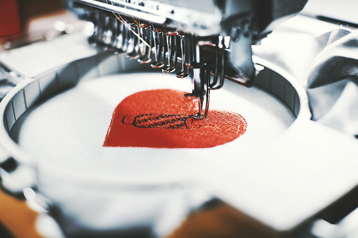 Close-up of an industrial embroidery machine stitching a red heart design with black detailing onto white fabric.